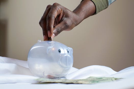 Close-up of a person's hand placing coins into a transparent piggy bank to save money.